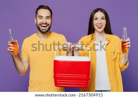 Young smiling fun cool couple two friends family man woman together wear yellow clothes hold red box freezer cooler refrigerator bottle drink beer isolated on plain violet background studio portrait