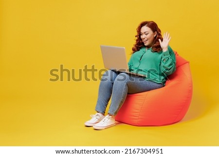 Full size body length young ginger chubby woman 20s wears green shirt sit in bag chair hold use work on laptop pc computer get video call wave hand isolated on plain yellow background studio portrait