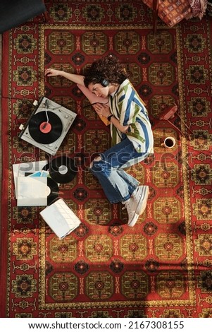 Above view of young woman in jeans and shirt looking at record player with vynil disk while listening to music in headphones on the floor Royalty-Free Stock Photo #2167308155