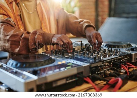 Hands of modern black man regulating sounds on dj set while standing by table in studio or loft apartment and turning mixers Royalty-Free Stock Photo #2167308369