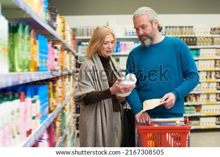 Aged blond female holding white plastic bottle with shampoo while showing it to her husband in department of bodycare products