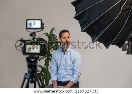 Behind the Scenes of Man Sitting on Chair on Video Production Set in Front of Video Camera, Tripod and Lighting Soft box. Ready For an Interview.