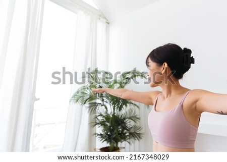 Young smiling attractive sporty Asian woman practicing yoga, doing Virabhadrasana 2 exercise, meditating in Warrior Two yoga pose, indoor working out at home, wearing sportswear. Half-length photo.