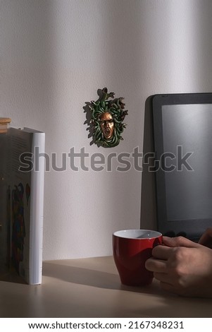 Close-up shot of a female hand holding a red cup. The statuette of an ancient Greek Gorgon Medusa framed by a ball of snakes is located on a wall. There is a monitor and books on the table.           