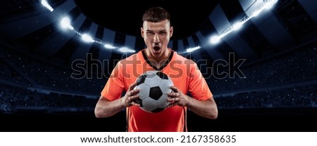 Winner emotions. Excited professional soccer, football player in football kit standing with ball and shouting over dark night stadium with flashlights background. Sport, competition, championship, wow