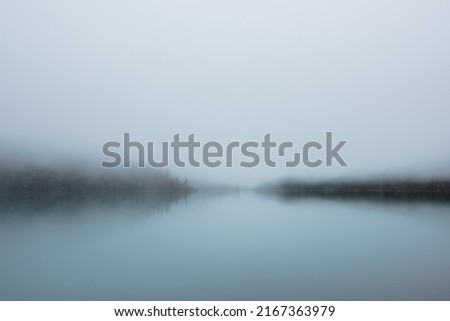 Tranquil meditative misty scenery of glacial lake with pointy fir tops reflection at early morning. Graphic EQ of spruce silhouettes on calm alpine lake horizon in mystery fog. Ghostly mountain lake. Royalty-Free Stock Photo #2167363979