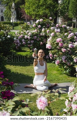 cheerful woman holding sports bottle while sitting in shoelace pose on lawn near blossoming peonies