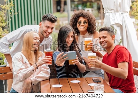 Group of happy young diverse friends, men and women, taking group selfie, while having a beer at the pub table outdoors