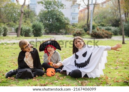 Treat or trick! Outdoor shot of three kids wearing carnival costumes for Halloween eating candies sitting in the park. Holiday celebration and Halloween traditions concept.