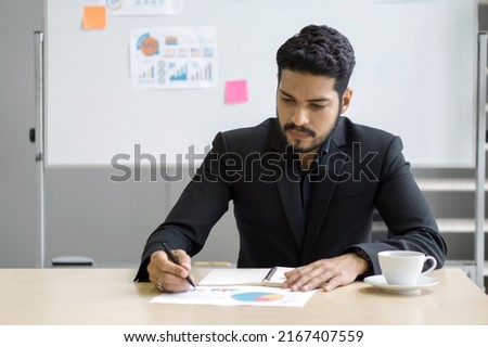 Young wavy hair, moustache and beard businessman in black suit writing on paper with pie chart. Calculate the best return on investment. There was a note paper and document on whiteboard.