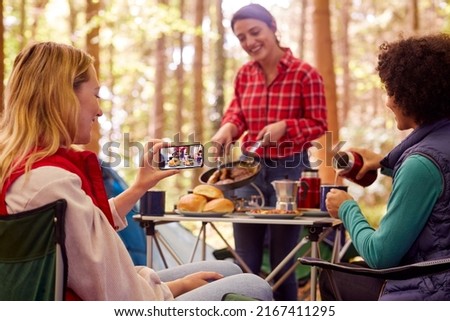 Woman Taking Photo Of Food As Female Friends On Camping Holiday In Forest Eat Meal Sitting By Tent