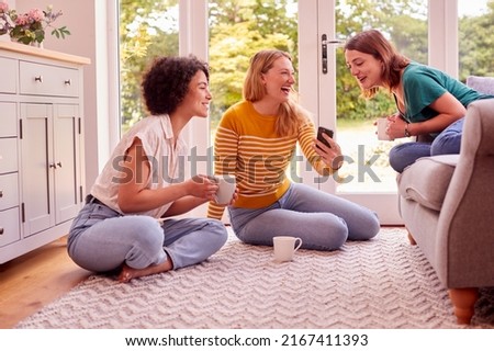 Group Of Female Friends Relaxing At On Sofa At Home Looking At Mobile Phone And Drinking Coffee