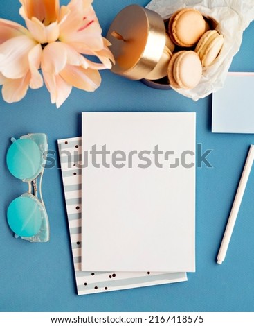 Home office workspace for freelance girl from above. Stationery flat lay with notebooks, pen, vintage sunglasses, pink flowers and macaroons on blue background. Blank dairy, drawing, notes template.