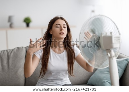 Sad young european woman suffers from unbearably too hot weather, catches cold air from fan in living room interior. Home without air conditioning, lady tired from summer heat, waves arms to cool down Royalty-Free Stock Photo #2167421781