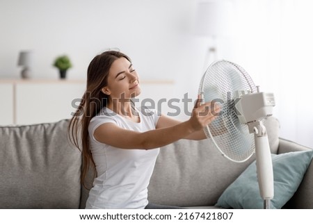 Glad young european woman suffers from heat sits on sofa catches cold air, turns fan to herself, enjoys cool down in living room interior. Summer weather, overheating without air conditioning at home Royalty-Free Stock Photo #2167421807