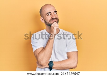 Young bald man wearing casual white t shirt with hand on chin thinking about question, pensive expression. smiling and thoughtful face. doubt concept.  Royalty-Free Stock Photo #2167429547
