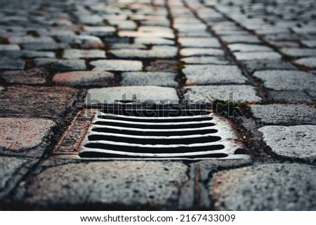 Grilled sewer cap on a sidewalk paved with cobblestone. Shot in perspective