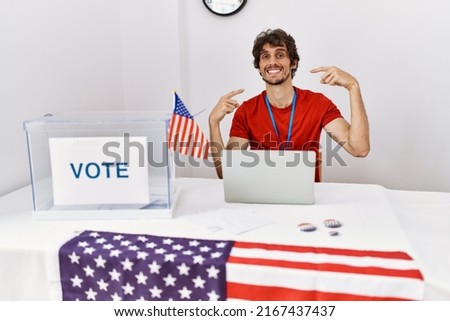 Young hispanic man at political election sitting by ballot smiling cheerful showing and pointing with fingers teeth and mouth. dental health concept. 