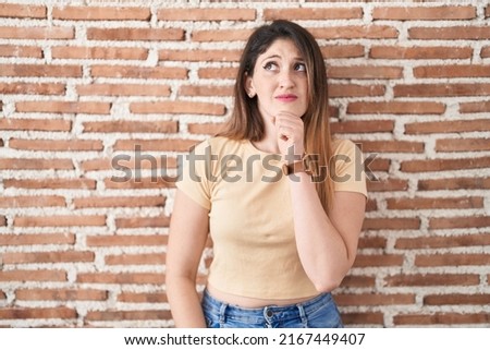 Young brunette woman standing over bricks wall thinking worried about a question, concerned and nervous with hand on chin 