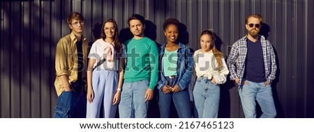 Handsome men and beautiful women in modern comfortable everyday clothes. Group portrait of six diverse relaxed young people in shirts, tees and jumpers leaning on street wall. Casual fashion concept Royalty-Free Stock Photo #2167465123