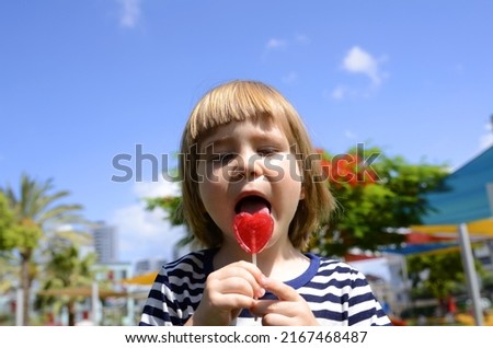 Portrait of a cute boy on the background of a bright playground. The child eats a heart-shaped candy. Flowering delonix regia. Concept: amusement park, family resort, traveling with children. sailing