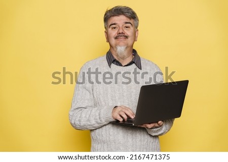 Mature man with grey beard smiling and using laptop isolated over yellow background