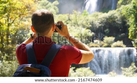 Young man blogger, zoomer traveler make photo for social media of amazing waterfalls, nature on trendy retro camera. Wanderlust, travel concept. Happy male tourist in trip, journey. Cinematic shot.
