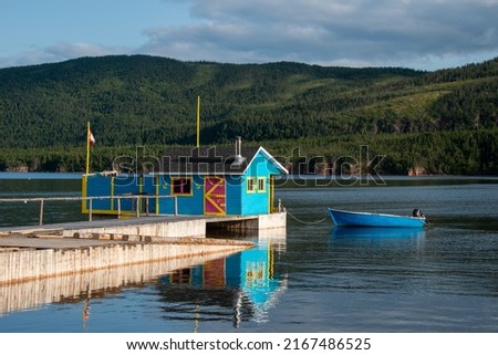 A small boathouse at the end of a wooden pier. The bright teal blue building has a red door and yellow trim. There's a small rowboat anchored to the wharf on a pond with a hill in the background. Royalty-Free Stock Photo #2167486525