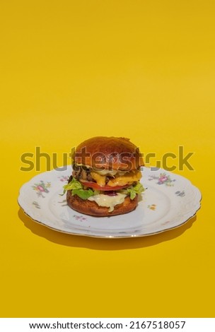 A juicy burger with meatball, cheese and fresh vegetables lies on a plate on a yellow background. Melting cheese flows down a bright and colorful burger. Restaurant burger. Delicious dinner