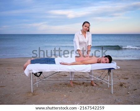 A young therapist performing a chiromassage treatment on a young man to treat a lumbar injury outdoors both looking at camera on a beach in Valencia. Royalty-Free Stock Photo #2167542259