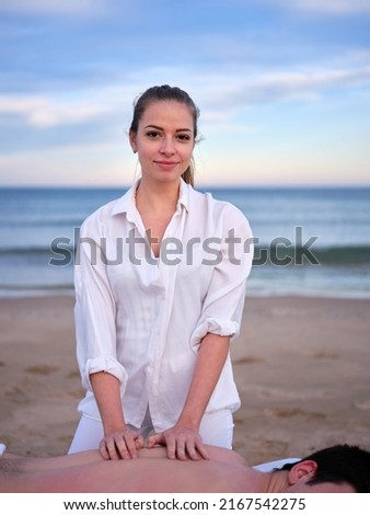 A young therapist performing a chiromassage treatment on a young man while looking at camera outdoors on a beach in Valencia. Royalty-Free Stock Photo #2167542275