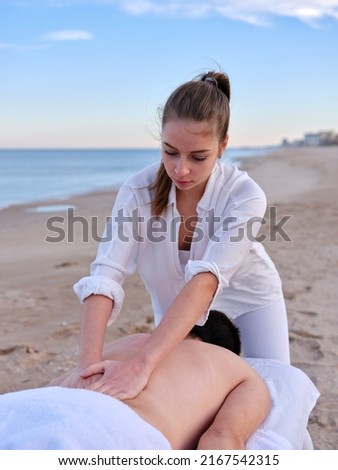 A young therapist performing a chiromassage treatment on a young man to treat a lumbar injury outdoors on a beach in Valencia. Royalty-Free Stock Photo #2167542315
