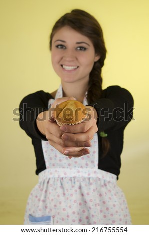 Smiling beautiful young hispanic brunette baker cook chef girl offering muffin to the camera over yellow background