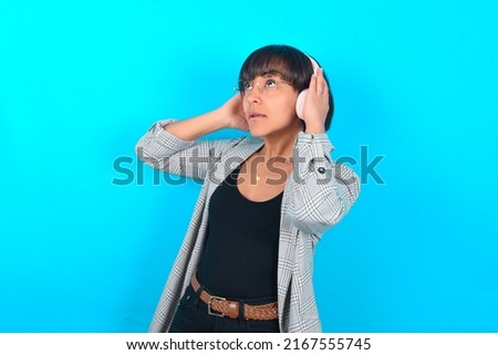 Young businesswoman with bob haircut wearing blazer against blue wall wears stereo headphones listens music concentrated aside. People hobby lifestyle concept