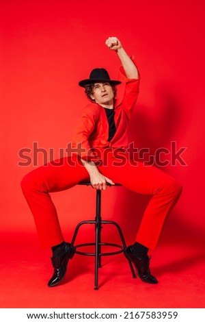 Portrait of a tall handsome man dressed in red shirt, red trousers and black hat posing with chair on the red background