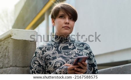 Young brown-haired Caucasian woman leaning against the wall waiting and holding her phone in her hand.
