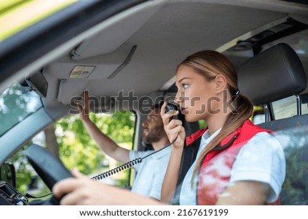 Young woman answering a call in an ambulance. She's with her colleague. They are wearing protective paramedic uniforms Royalty-Free Stock Photo #2167619199