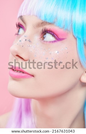 Anime makeup. Pretty girl with bright makeup, glitter freckles and in colored violet-blue wig looks away. Pink background. Hairstyle, hair coloring, make-up. Japanese anime style. 