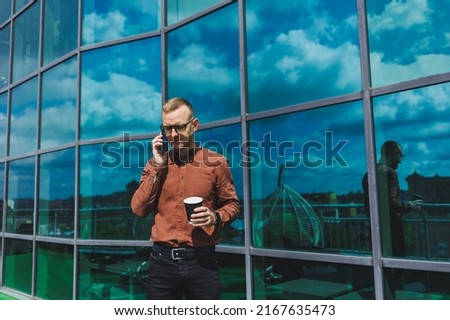 Attractive fashionable male businessman in a stylish shirt and trousers talking on the phone near the office building and drinking coffee, modern glass office