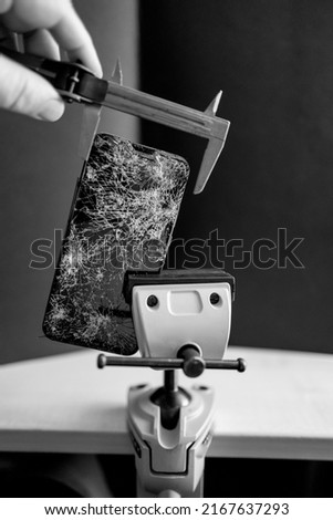 a completely destroyed cell phone is clamped in a vice and measured with a gauge