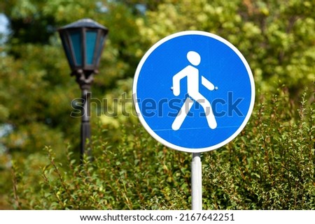 pedestrian zone road sign close-up on a background of park green trees. round blue road sign with white human icon. pedestrian zone in the park road sign next to a vintage street lamp. Royalty-Free Stock Photo #2167642251