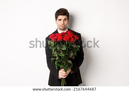 Image of handsome boyfriend in black suit, holding bouquet of red roses and smiling, being on a date, standing over white background