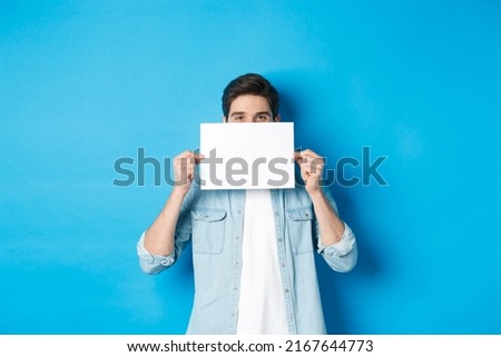 Sneeky handsome guy hiding face behind blank piece of paper for your logo, making announcement or showing promo offer, standing over blue background