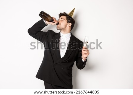 Image of handsome guy in trendy suit, drinking champagne from bottle and celebrating holiday, having a birthday party, getting drunk while standing over white background