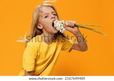 funny, cute girl posing standing on a bright background in yellow clothes and with a bouquet of daisies in her hands, trying to bite them. Horizontal studio photography