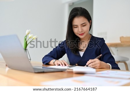 Beautiful elegant young asian businesswoman or female manager in formal suit sits at her office desk, reviewing and signing a document.