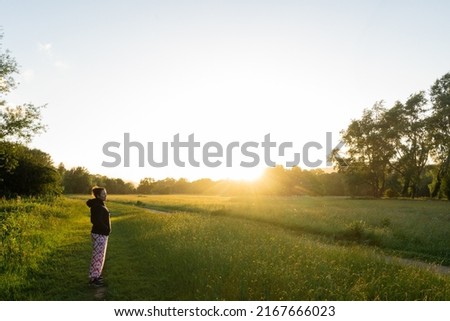 A young lady in a meadow in the British countryside gazing across the grassy field towards the setting sun. the are has a footpath and is popular with ramblers and hikers Royalty-Free Stock Photo #2167666023