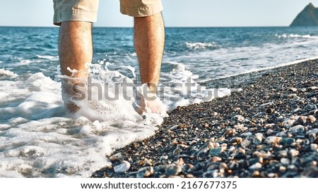 Man wearing shorts, walking barefoot along the seashore. Male legs walks on pebble beach along the shore near the water with waves, low section. Wellness, freedom and travel in summertime concept. Royalty-Free Stock Photo #2167677375
