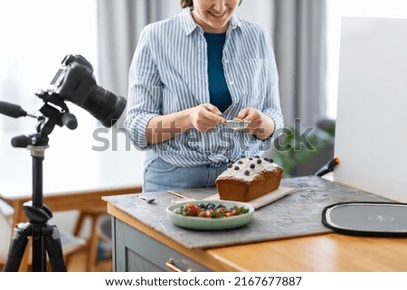 blogging, photographing and people concept - happy smiling female photographer or food blogger with camera pouring powdered sugar to cake in kitchen at home