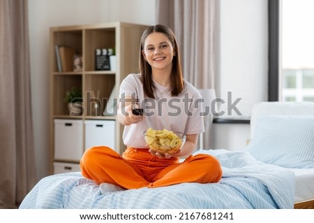 unhealthy eating, fast food and people concept - happy girl with remote control and crisps watching tv sitting on bed at home